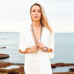 Woman with fair skin, blonde hair and a white dress, looking at the camera wearing Ambarya Wisdom - Lapis Lazuli Mala Bead Set. She is holding onto the necklace that is around her neck